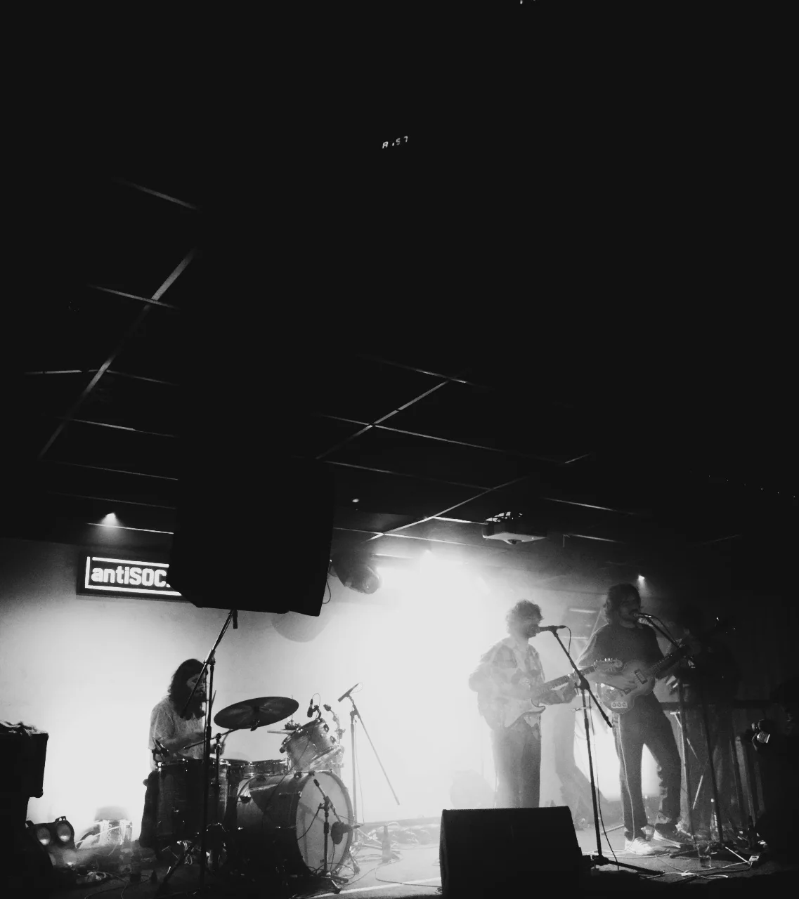 A black and white photograph of a drummer and guitarists performing on a brightly backlit indoor stage.