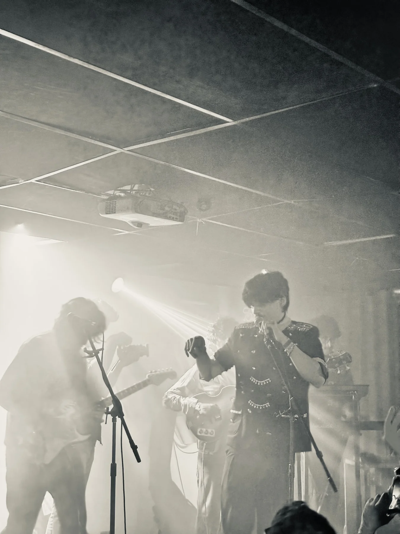 A singer gesturing beside a microphone with bandmates playing guitars in soft stage haze.