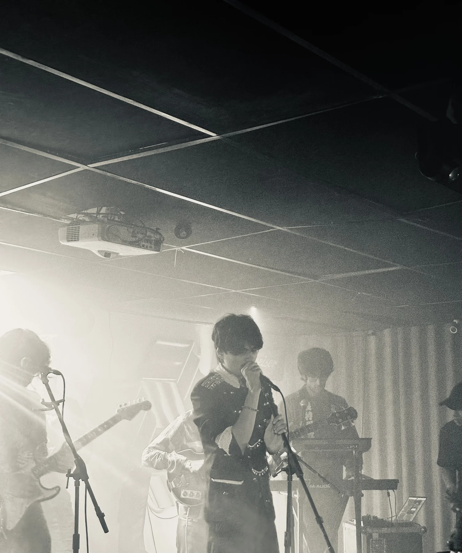 A singer at a microphone with guitarists on an indoor stage under hazy white lighting.