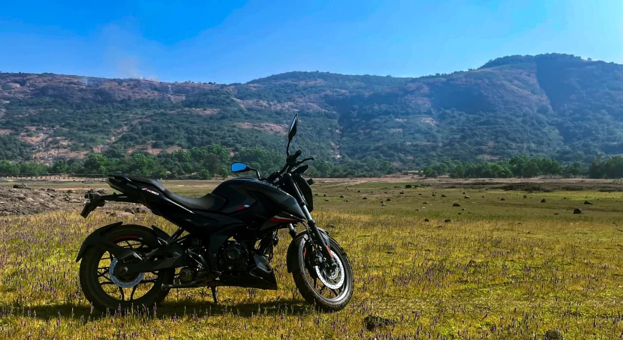 A black motorcycle parked in a field of small purple flowers beneath a rugged green hillside and blue sky.